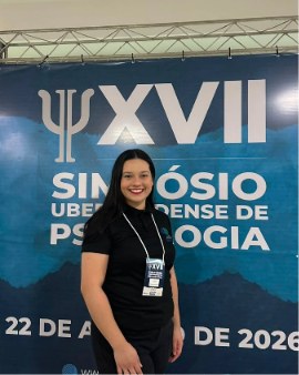 Karine sorrindo, de crachá, diante do painel do XVII Simpósio Uberlandense de Psicologia