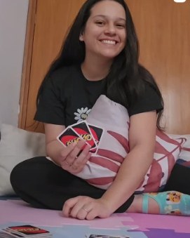 Karine sorrindo, sentada no chão abraçada a uma almofada, com cartas de um jogo nas mãos